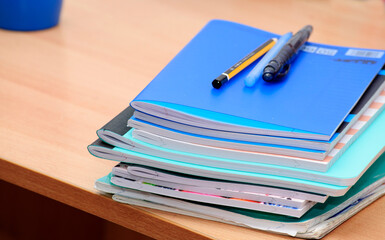 Notebooks in blue, pink and other colors, pens and a pencil on a wooden desk. Close-up photo.