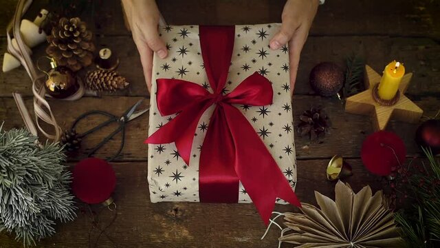 Hands decorating stylish christmas gift with green fir branch under red ribbon on rustic wooden table with festive holiday decorations. Top view. Happy Holidays! Atmospheric Xmas footage