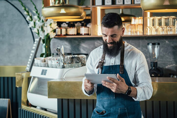 Handsome Barista Using Digital Tablet in a Cafe.

Smiling waiter with a beard leaning on the bar...