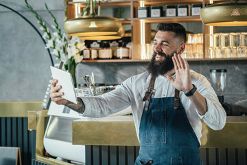 Happy Barista Using Digital Tablet for Talking on Video Call in a Cafe.
Cheerful smiling waiter saying hello or goodbye and waving at tablet screen to family or friends while working in a coffee shop.