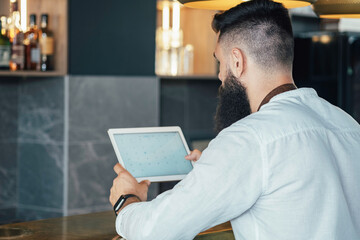 An Anonymous Barista Using Digital Tablet in a Cafe. 
Unrecognizable waiter with a beard leaning on the bar counter and reading online order or watching something on a digital tablet while working.