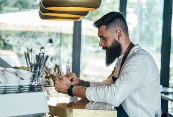 Handsome Barista Using Digital Tablet in a Cafe.
 
Serious bartender with a beard standing on the...