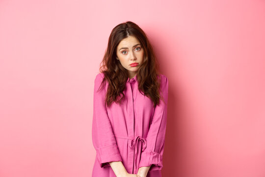 Cute coy girl making silly face, wants something, begging for gift, standing against pink background