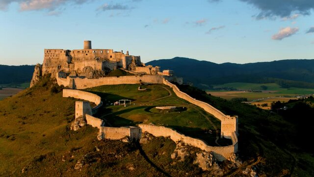 Aerial view of Spi&scaron; Castle lit by orange setting sun. Extensive castle ruins, standing on a 200m high white rock above the valley against mountains in background. UNESCO site. Slovakia.