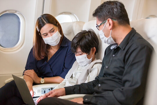 Asian family talking while sitting on seat in an airplane. Father, mother and son sitting and using computer laptop on airplane cabin. Asian passenger wearing medical face mask during travel abroad