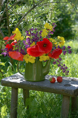 Still life in a sunny summer garden with a cheerful red-yellow-purple bouquet of flowers in a green teapot on a small wooden bench in rustic style.