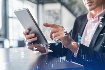 Businessman in suit using tablet device to optimize trading strategy at corporate finance fund. Forex chart hologram over modern panoramic office background