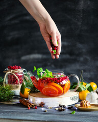 Winter drink on a dark background. Cranberry tea with tangerines and mint. A woman's hand in food photography. A warming berry drink is being prepared. Put the berries in the kettle.Hot tea with honey