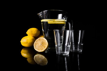 Jug or pitcher of water with lemons and two empty glasses, on black background.