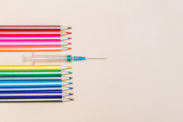 An empty syringe with a needle lies among colored pencils