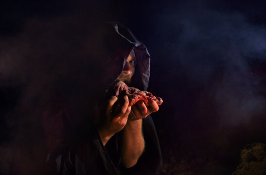 A crazy man in a black hood eats raw meat in a dark street