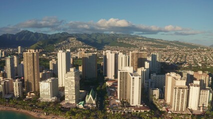 Naklejka premium Panning aerial drone slowly flying over a colorful Honolulu Skyline while Sunset in Oahu, Hawaii with Waikiki Beach Shooting from a bird's eye view. Magnificent mountains of Hawaiian island of Oahu.