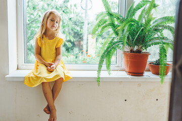 5 years old blonde girl sit on the windowsill with sad face. Quarantine, lockdown. Stay at home.