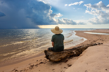 Fototapeta na wymiar Man in hat sit on tree trunk looking at storm and sunset on Tapajos river beach in Amazon Rainforest. Concept of nature, conservation, environment, ecology, vacation, tourism, travel. Alter do Chao.