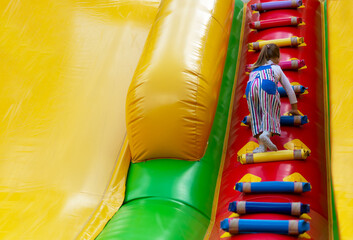 Fototapeta na wymiar A little girl is having fun on a high inflatable slide and climbs up the stairs. A small child is having fun on an inflatable trampoline. A happy, fun childhood. Entertainment for children. Play time