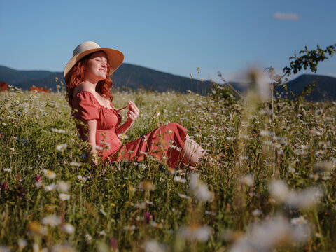 red-haired woman wearing dress and hat on summer evening field of daisies reading a book while sitting on a blanket . concept of digital detox, digital cleanse, reconnecting with nature.