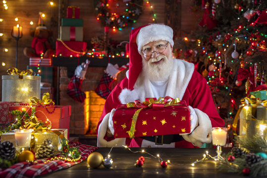 Santa Claus with gift box presents near fireplace and Christmas tree. Wooden house, New Year's cheerful mood Spirit of Christmas. Senior man with real white beard cosplay Father Christmas.