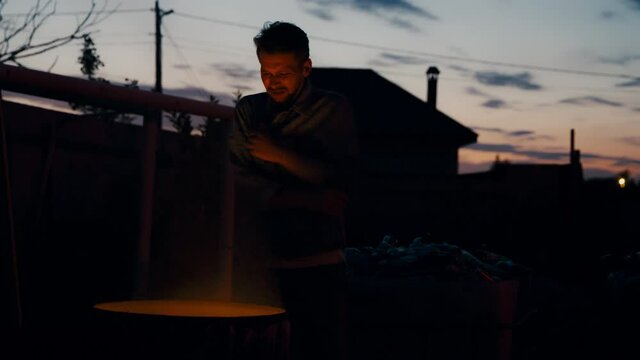 A European man in jean jacket near a burning garbage can on his face.