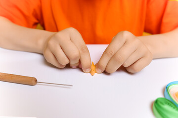 a child in an orange T-shirt glues paper thin strips for applications in the technique of quilling