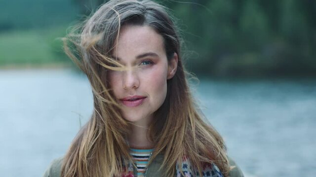 portrait independent young woman looking confident with serious expression running hand through hair with wind blowing outdoors by lake 4k