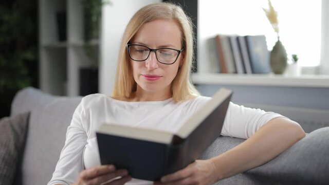 Pretty caucasian woman in eyeglasses and casual outfit reading book during free time at home. Mature blonde sitting on couch and enjoying new fascinating novel.