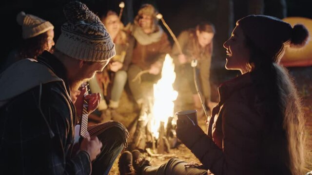 friends sitting by campfire young man playing ukulele singing to woman enjoying romantic serenade camping in forest at night