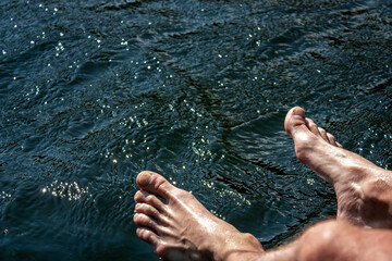 Tanned feet of a man against a background of blue water, top view. Two feet above the water. The sun illuminates the feet of a person and the water of the river.