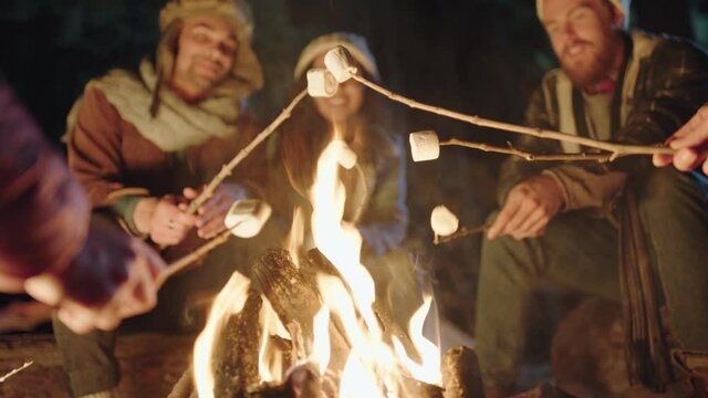 group of friends sitting by campfire roasting marshmallows chatting sharing stories camping in forest at night enjoying outdoor adventure
