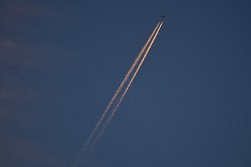 Beautiful telephoto evening view of airplane contrail in orange colors of setting Sun on dark blue background seen over Dublin Mountains, Ireland. Golden hour minimalism concept