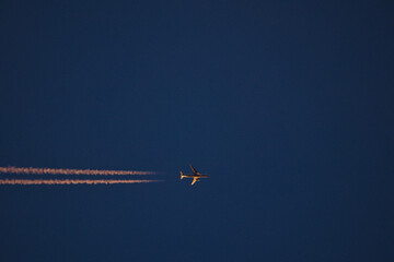 Beautiful telephoto evening view of airplane contrail in orange colors of setting Sun on dark blue...