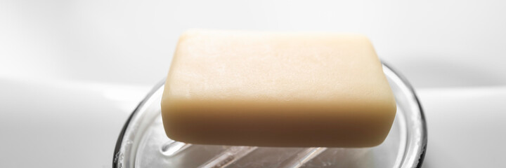 Beige-colored bar of soap on the transparent saucer. Soap bar isolated on white bathtub backdrop with space for text and design.