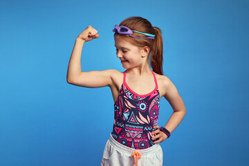 Strong kid girl spends raises arm and looking at muscles, ready for swim, poses over blue...
