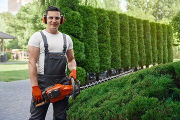 Obraz premium Front view of competent gardener in brown overalls, safety glasses and gloves posing at garden with electric trimmer in hands. Seasonal work at summer garden. 