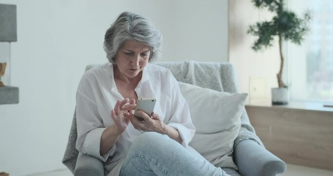A happy old adult lady spending time at home using her cell phone and having fun communicating with her kids and friends in social media. Receiving a text message from her daughter or son.