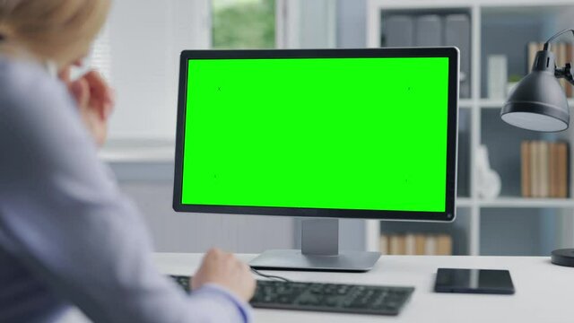 Mature woman with blond hair sitting at desk and typing on computer with chroma key screen. Female office manager using modern technology for work.