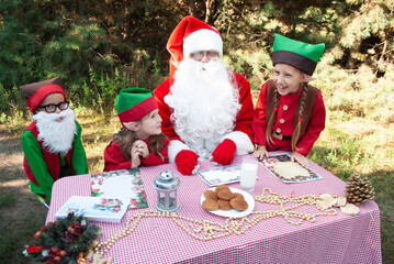 Santa and the elves sit in the woods against the background of Christmas trees at the table dismantling the mail. The concept of Christmas and New Year