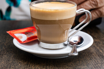 Close-up of a cup with latte, in a white mug, along with a teaspoon and an envelope of red sugar.