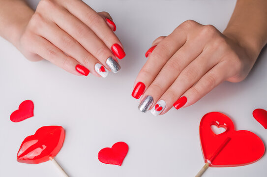 The hands of a young woman. The nails are covered with red gel polish for Valentine's Day. On a white background with lollipops in the shape of a heart and lips. Nail art and design ideas.