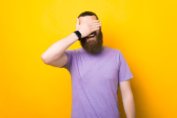 Smiling happy man is covering his eyes with his hand over yellow background.