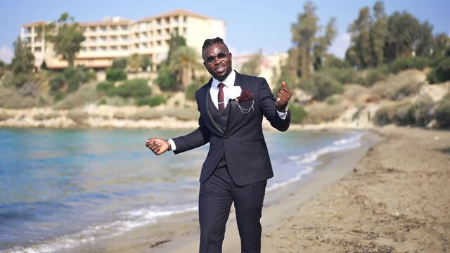 Portrait of cheerful excited happy African American groom in elegant suit dancing walking on sunny sandy Mediterranean sea beach smiling. Joyful handsome man having fun on wedding day outdoors
