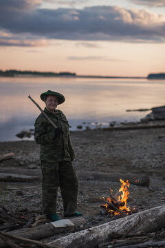 A young woman in camouflage clothes builds a fire on the bank of the river. Dusk time