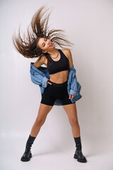 Cheerful young woman is dancing on a light background in a denim jacket and black sports shorts and boots. Caucasian brunette girl posing on background in studio