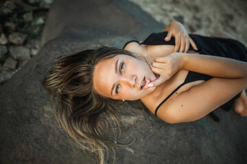 Beautiful young curvy woman in the black dress lying on stones at sunset. Wild nature, natural beauty