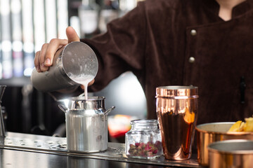 Barman gently pours finished cocktail from glass shaker into metalic glass. Body of bartender in brown apron on background.