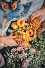 Happy family having a picnic with croissants and fruits decorated with flowers, pine branches and cones, close-up view of their hands with food