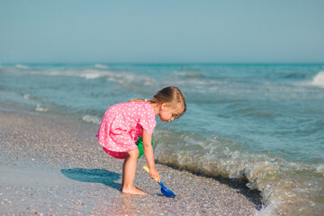 A little girl plays with a shoulder blade and a bucket on the beach in a pink heart dress