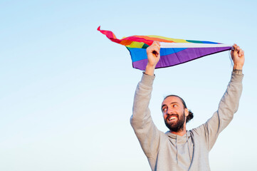 Cheerful boy with a lgbtq rainbow flag on the beach. Young man holding a rainbow flag against blue sky