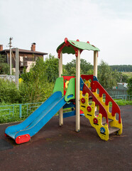 A wooden slide of bright red, blue, yellow color against the background of a village house and greenery on a clear sunny day. Playgrounds, sports, entertainment.
