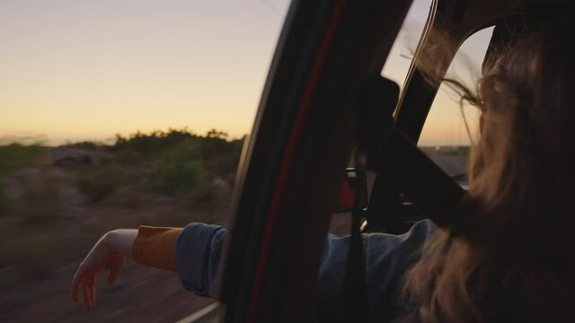 woman in car holding hand out window feeling wind blowing through fingers driving in countryside on road trip travelling for summer vacation enjoying freedom on the road at sunrise