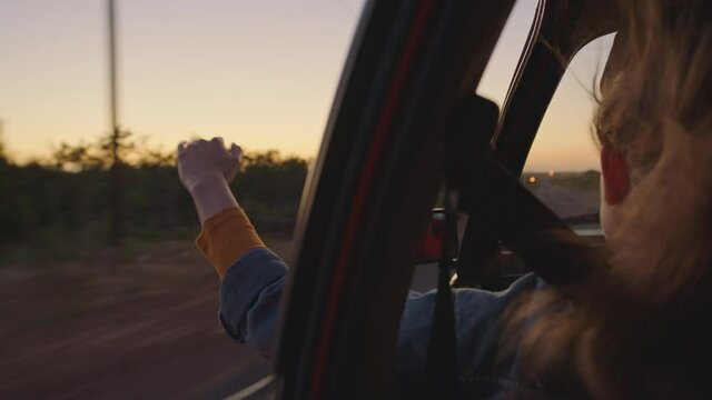 woman in car holding hand out window feeling wind blowing through fingers driving in countryside on road trip travelling for summer vacation enjoying freedom on the road at sunrise